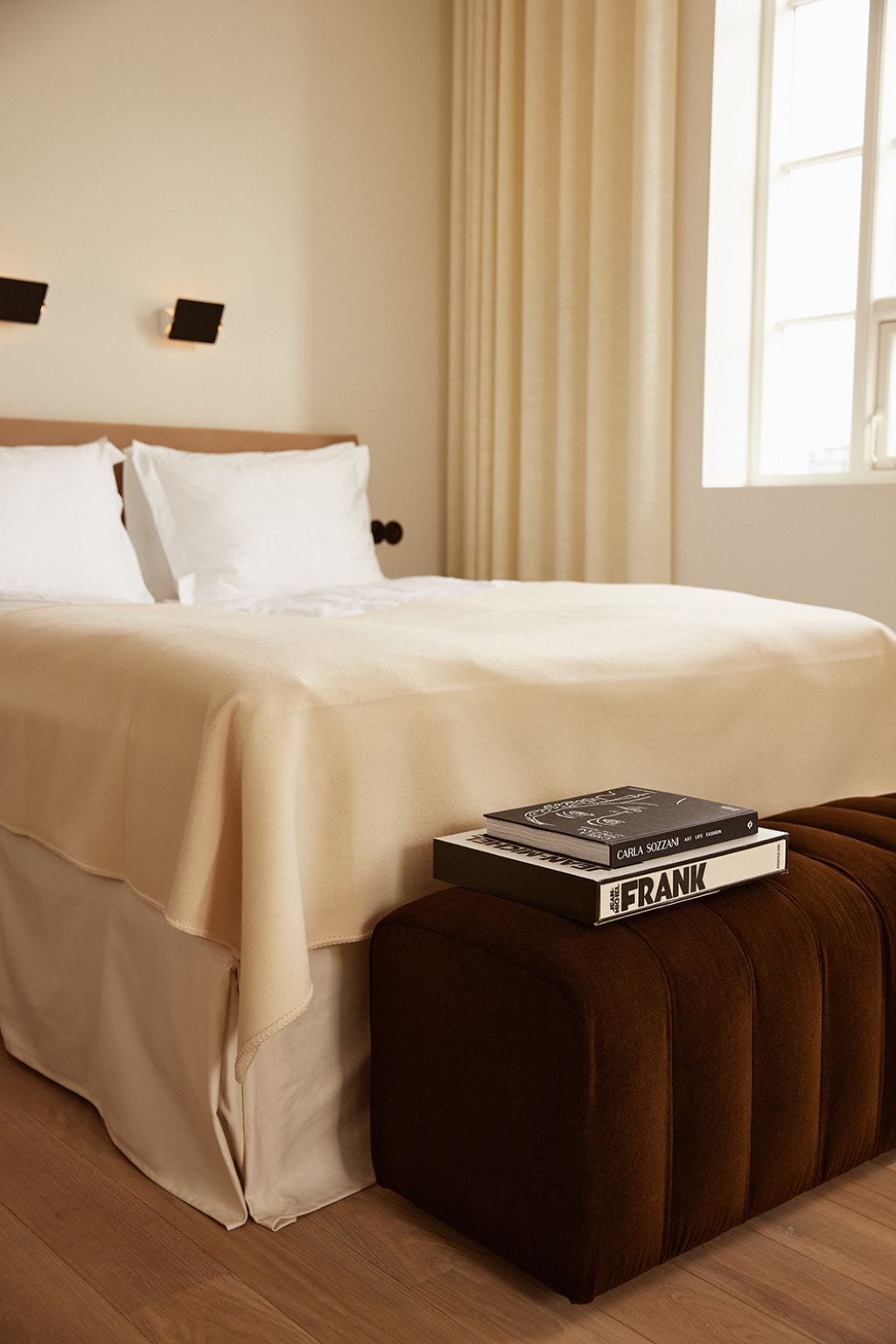 A neatly made bed with white linens, a brown ottoman, and two books on top.