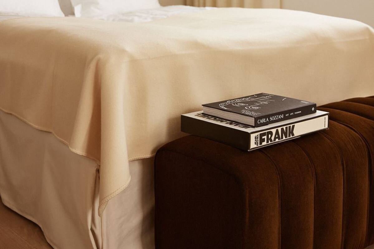 Cama bem arrumada com roupa de cama branca, um banquinho marrom e dois livros em cima.