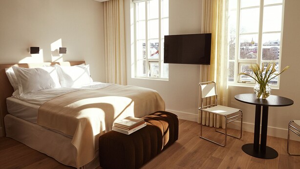 A hotel room with a bed, a TV, a small table with a vase, and a view of the city through large windows.