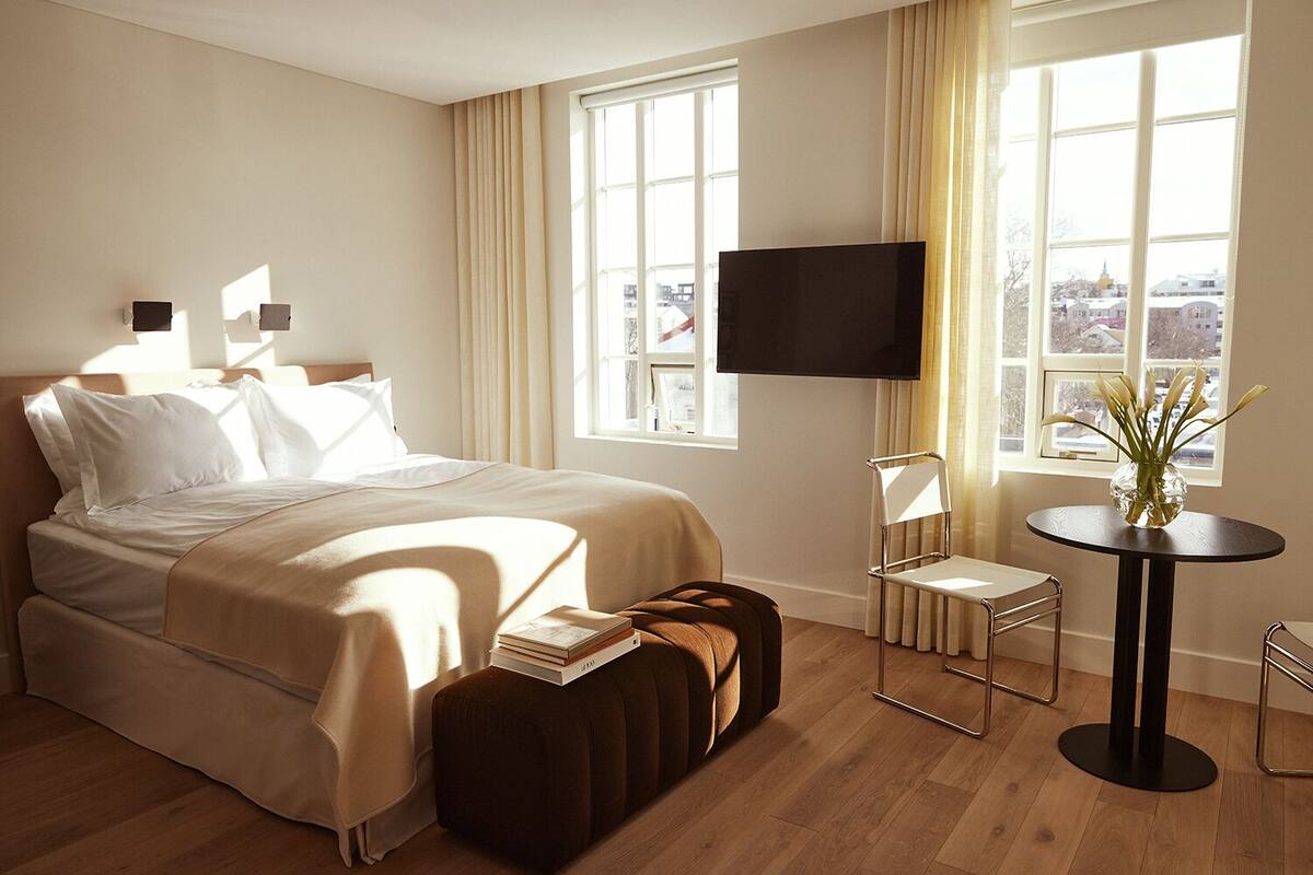 Quarto de hotel com uma cama, uma televisão, uma mesinha com um vaso e vista da cidade através de amplas janelas.