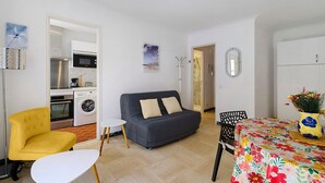 Ein Wohnzimmer mit einer Couch, einem gelben Sessel, einem kleinen Tisch, einem Esstisch mit einer blumigen Tischdecke und einer Küchenzeile mit Geräten.