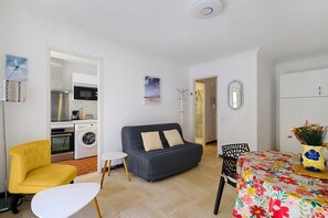 Ein Wohnzimmer mit einer Couch, einem gelben Sessel, einem kleinen Tisch, einem Esstisch mit einer blumigen Tischdecke und einer Küchenzeile mit Geräten.