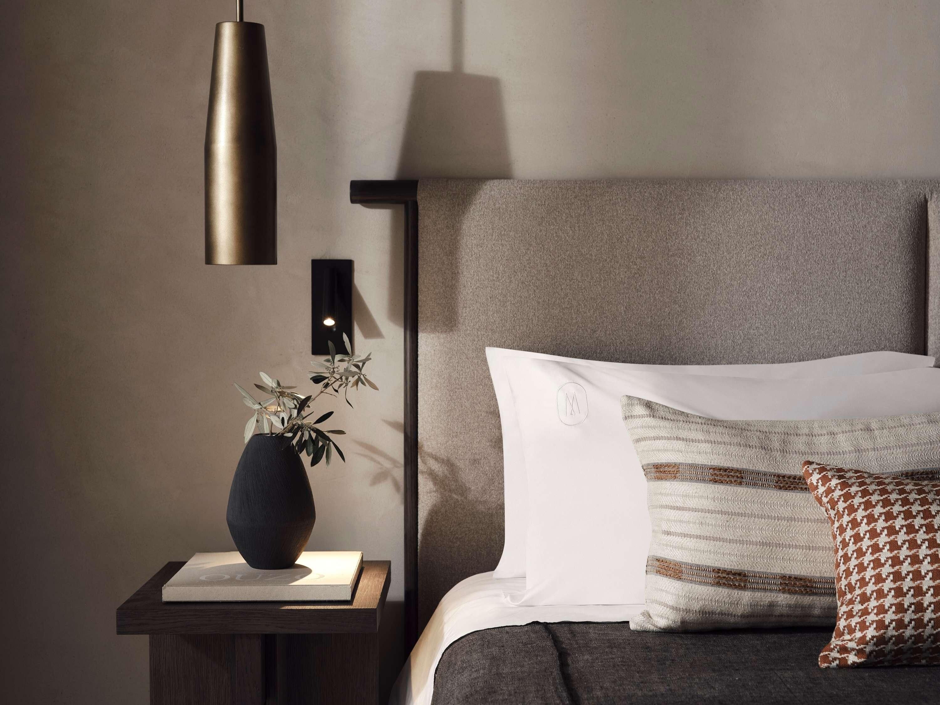Ein ordentlich eingerichtetes Schlafzimmer mit Nachttisch, Lampe, einer Pflanze in einer Vase, einem Bett mit Kissen und einer Wandleuchte.