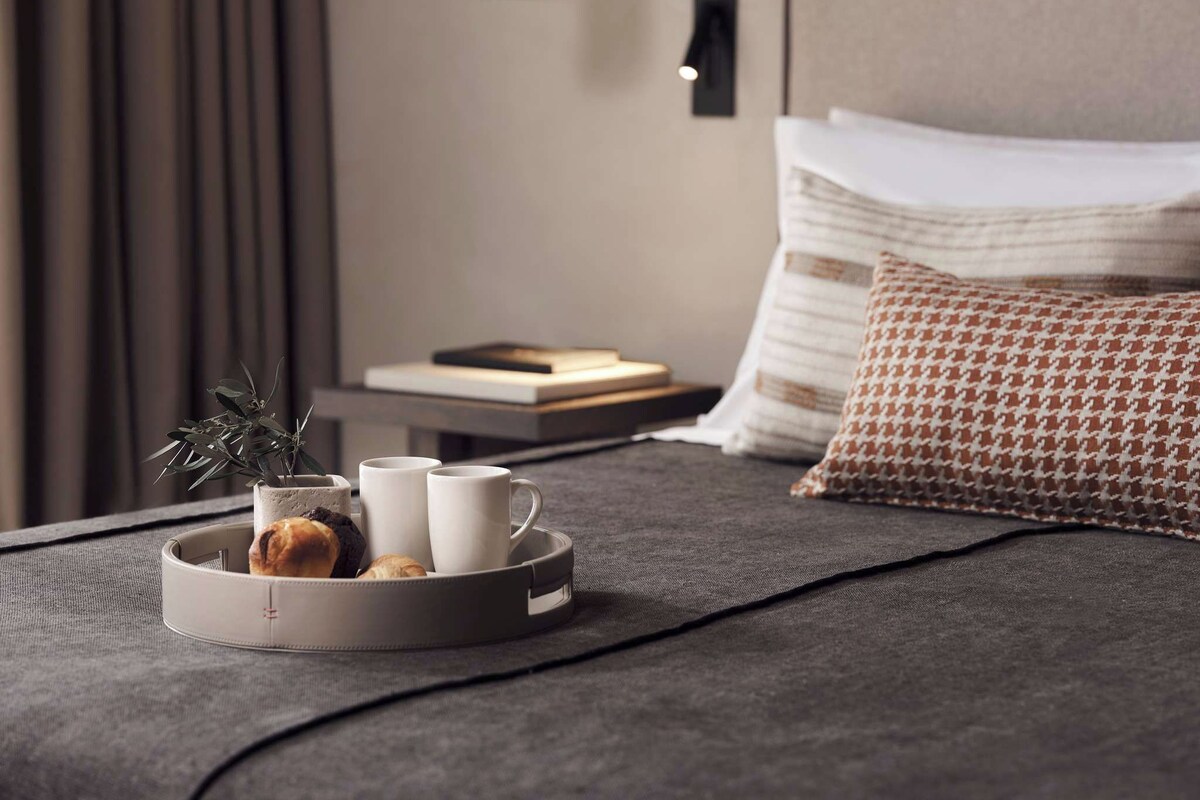 Una cama bien hecha con un edredón gris, una bandeja con pasteles y tazas, una planta y un libro en una mesita de noche.