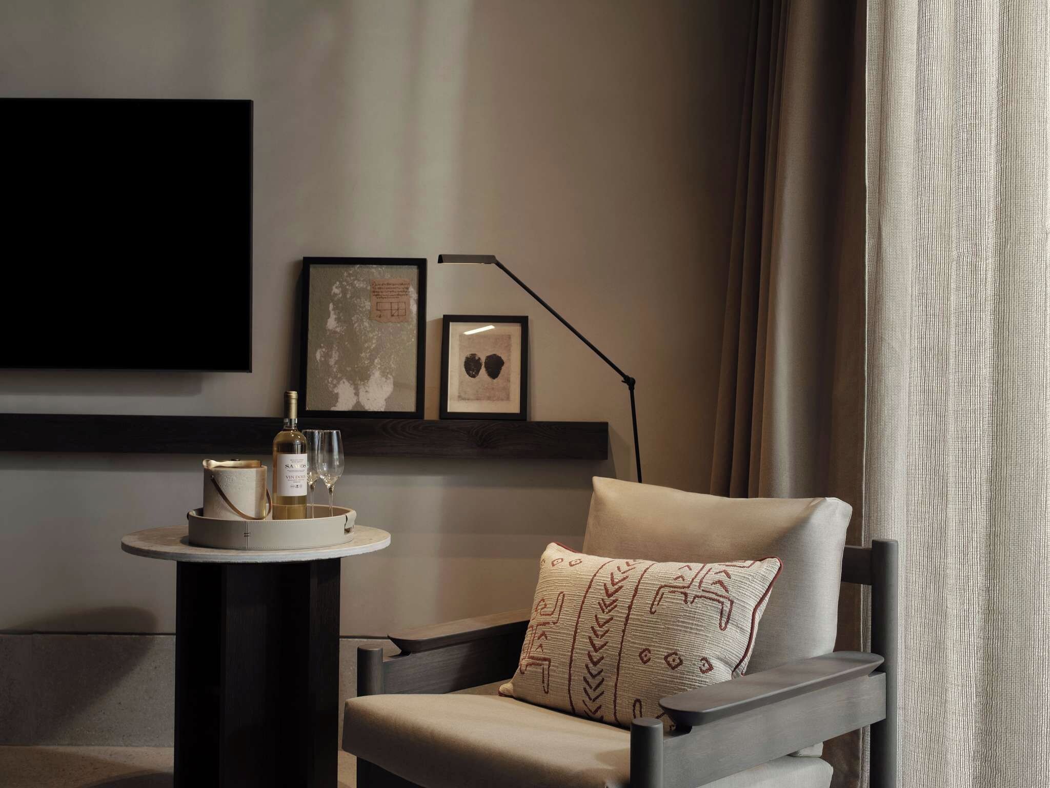 A modern room with a dark wooden chair, a round table with a bottle and glasses, and two framed pictures on the wall.