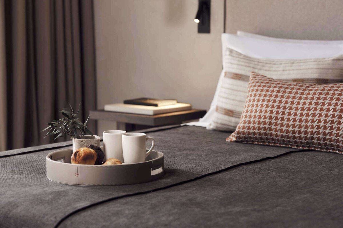 A neatly made bed with a grey blanket, a tray with pastries and cups, a plant, and a book on a bedside table.