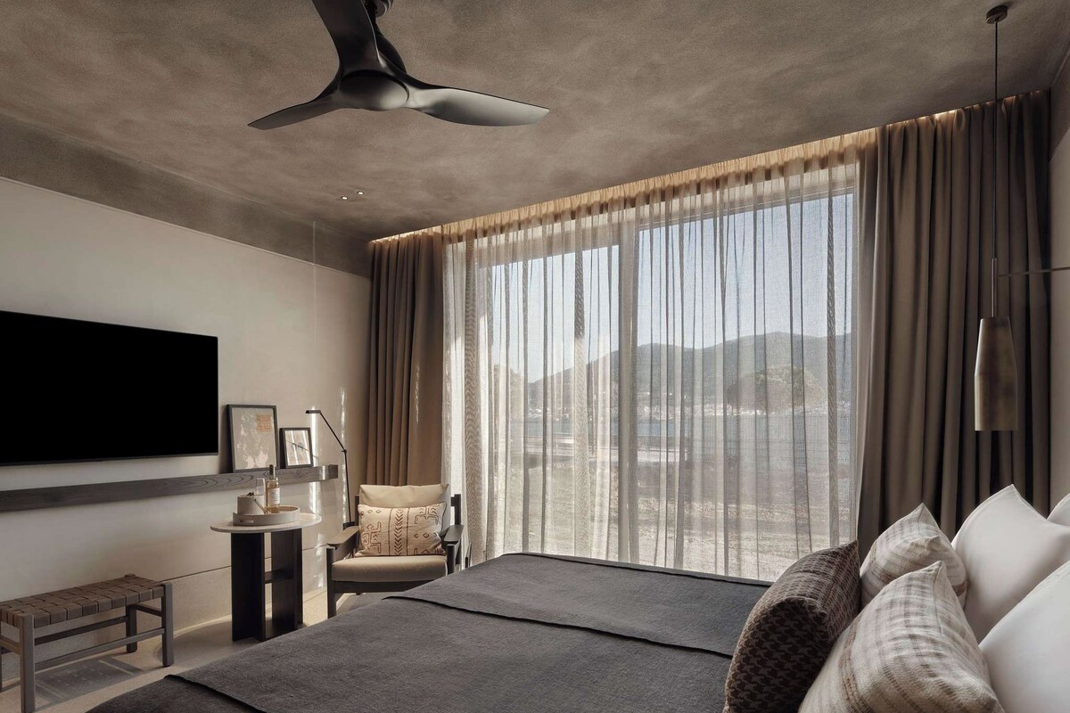 Un dormitorio moderno con una cama grande, una silla, una mesita pequeña y un televisor montado en la pared.