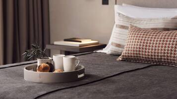 A neatly made bed with a grey bedspread, a tray with a plant and two mugs, a book, and a patterned pillow.