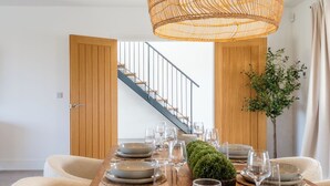 A dining area with a wooden table set for six, a hanging rattan light fixture, and a staircase in the background. - Luxury Cotswolds Estate with Pool (Wootton)