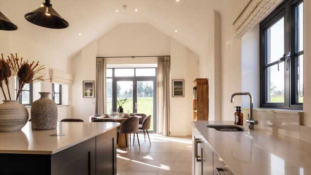 A modern kitchen with a large island, a dining area with chairs, and a view of the outdoors through a large window. - Luxury Cotswolds Estate with Pool (Wootton)