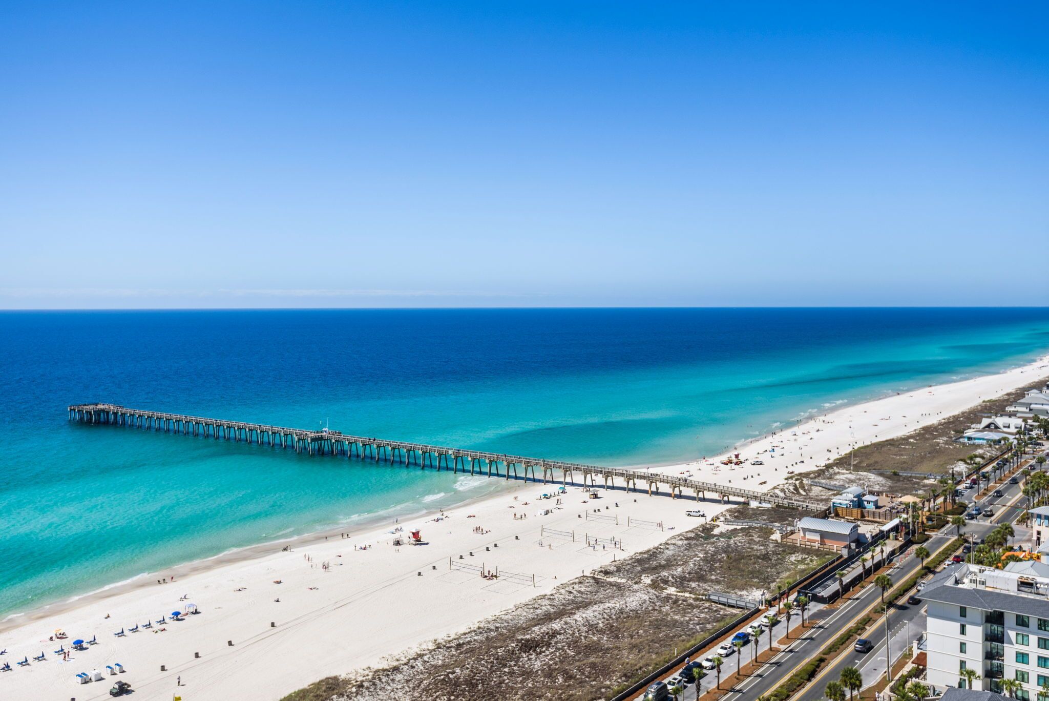 A beach with a pier extending into the ocean, a sandy beach, and a road parallel to the beach.