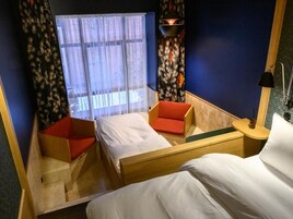 A hotel room with two beds, a small seating area, a window with sheer curtains, and a nightstand with a lamp.