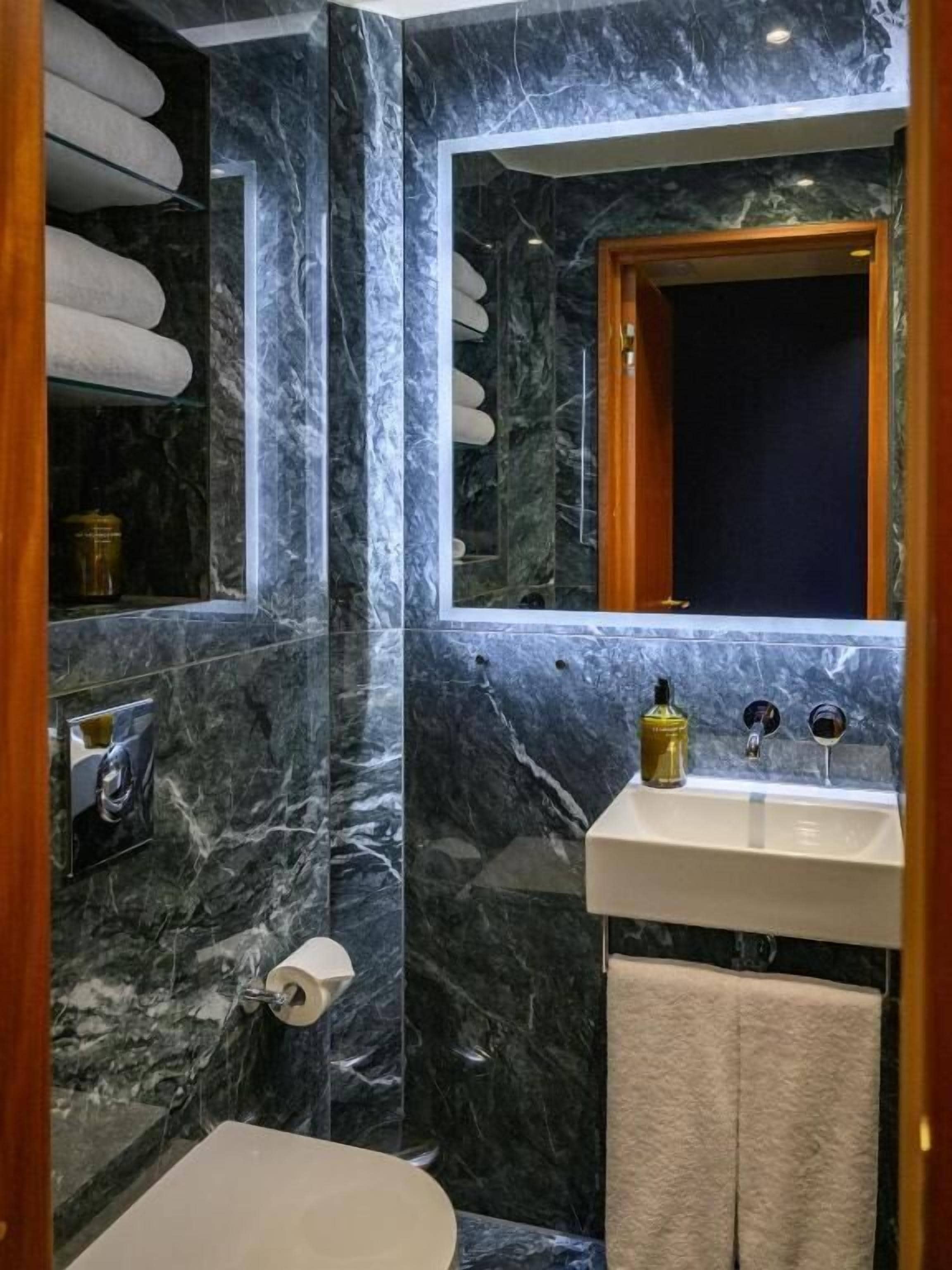 Un baño con paredes de mármol, un espejo con marco de madera, un lavamanos blanco y un perchero para toallas.