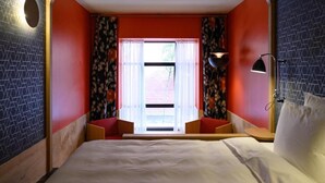 Une chambre d’hôtel avec un lit, une fenêtre avec des rideaux, une lampe fixée au mur et un petit coin salon.