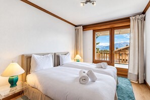 A hotel room with a bed, two bedside tables, a window with curtains, and a view of mountains. - Golf Park Duplex by Mrs Miggins – Elegant Apartment in the Heart of Crans-MontanaWelcome to Golf Par (Crans-Montana)