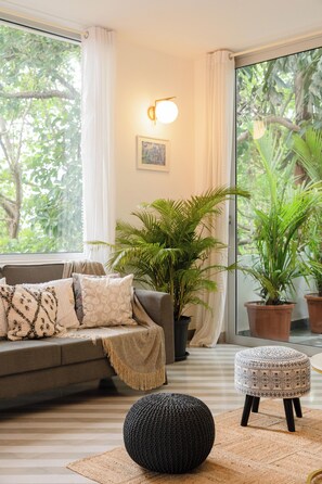 Een woonkamer met een bank, voetenbankje, potplanten en een wandlamp.