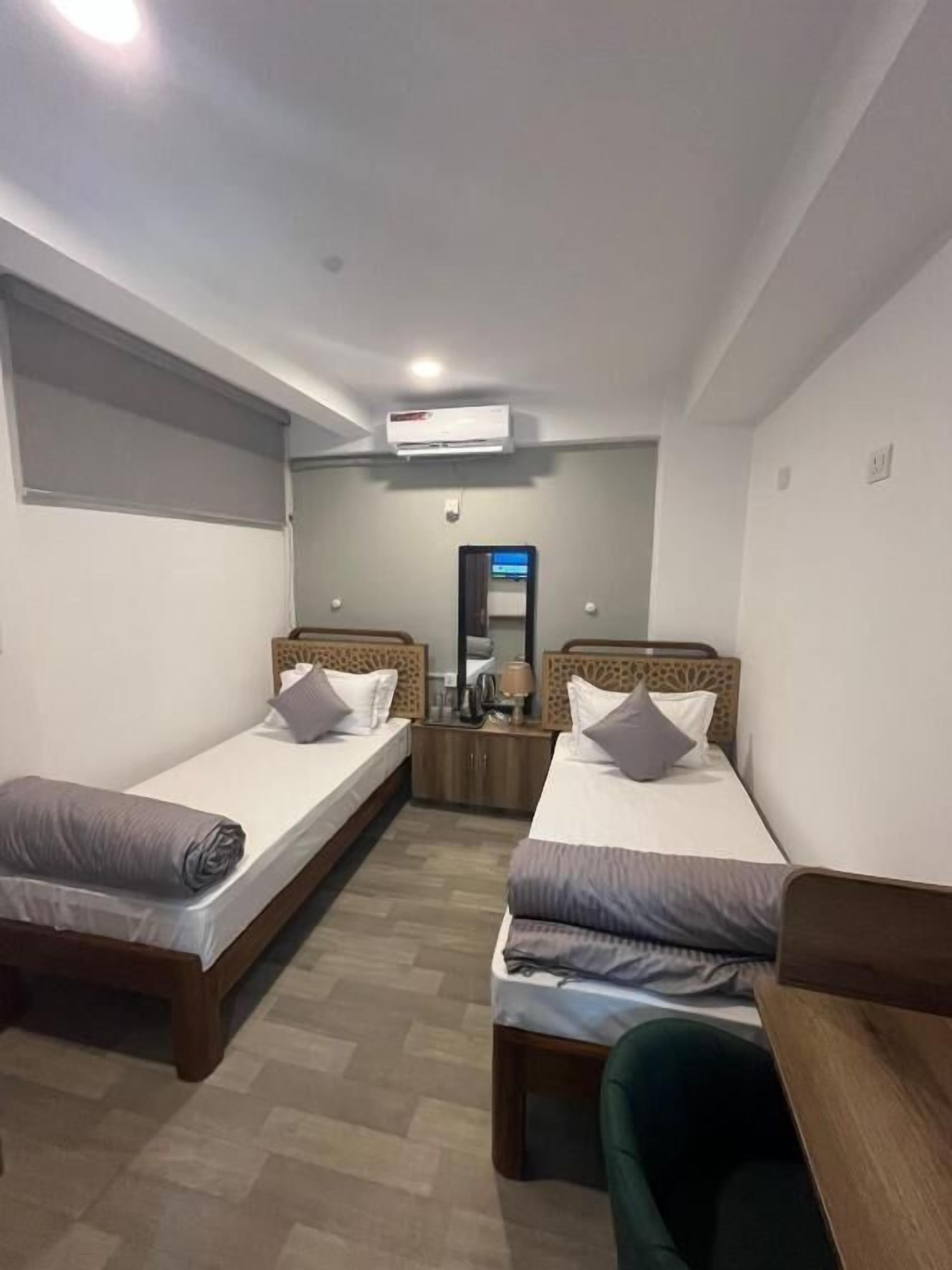 A hotel room with two single beds, a small desk, a chair, a mirror, and an air conditioning unit.