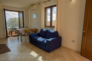 A living room with a sofa, a dining table with chairs, a window with curtains, and an air conditioning unit. - Appartamento Lido Residence 1 (Cala Gonone)