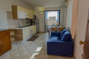 A compact kitchen with a dining area, a sofa, and a view through an open door. - Appartamento Lido Residence 1 (Cala Gonone)