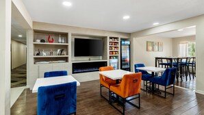 A modern interior with a flat-screen TV, seating area with blue and orange chairs, a refrigerator, and a dining table. - Sonesta Essential Hotels Longview (Longview)