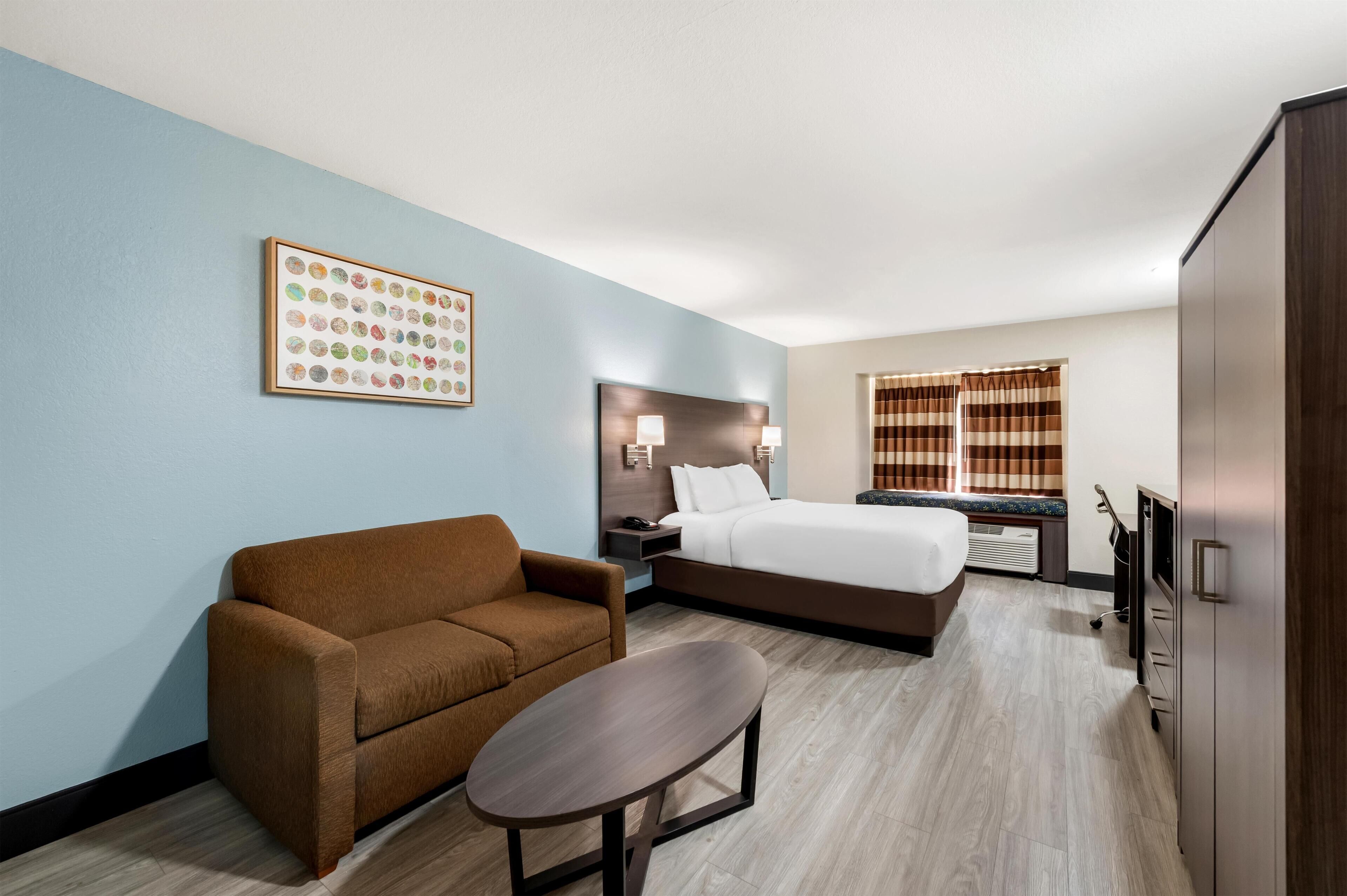 Une chambre d’hôtel comprenant un lit, un canapé, une petite table, une tête de lit avec une lampe, une œuvre d’art accrochée au mur et une fenêtre avec des rideaux.