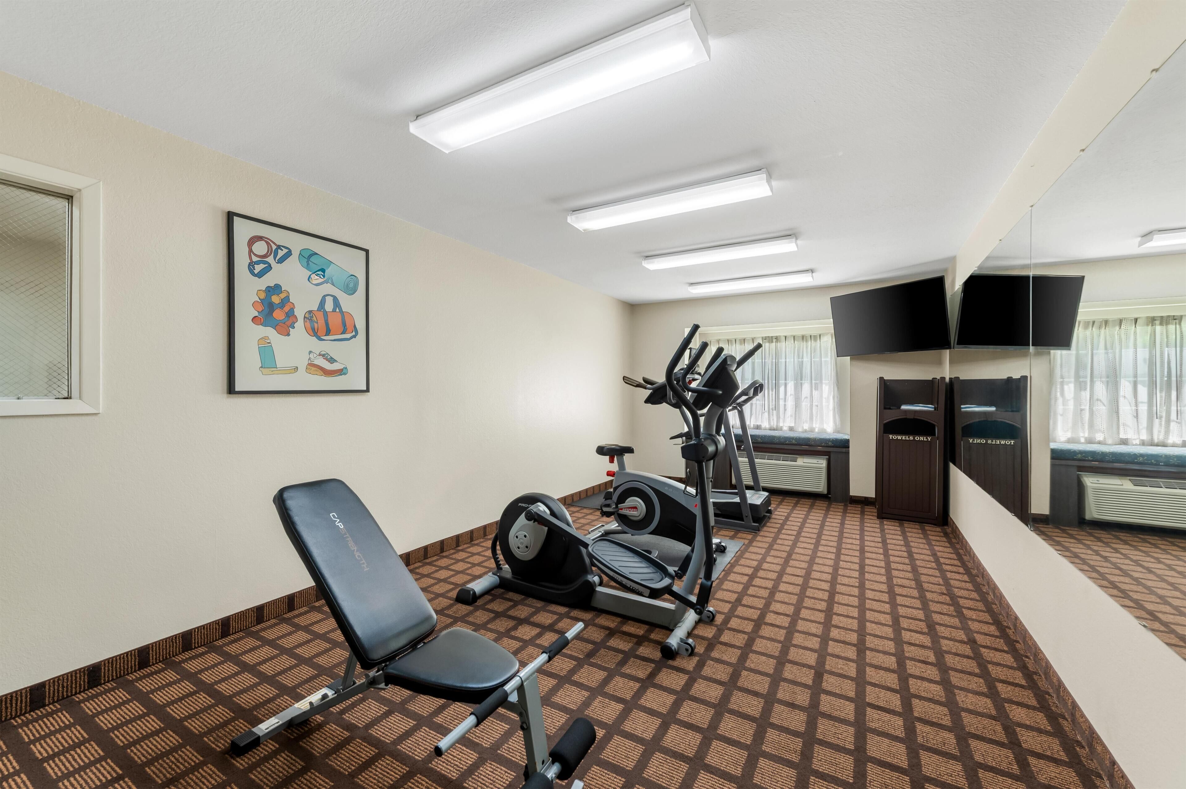 Gimnasio de hotel con equipo de ejercicio, una banca, un perchero para toallas y un televisor.