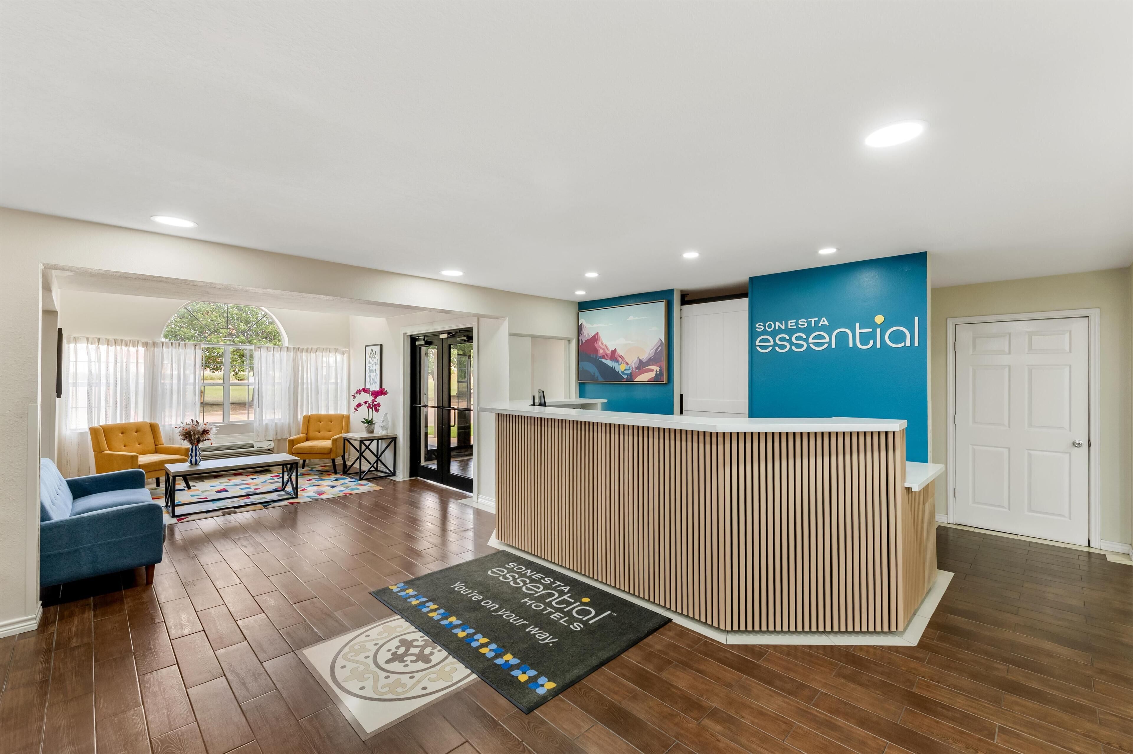 Un espace d’accueil d’hôtel comprenant un comptoir de réception en bois, un mur bleu orné du nom de l’hôtel, un tapis placé devant le comptoir, un canapé bleu, deux fauteuils jaunes et une table basse.