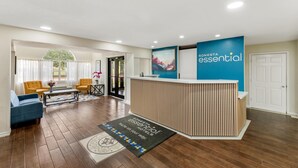 A hotel reception area with a wooden reception desk, a blue wall with the hotel name, a reception desk mat, a blue sofa, two yellow armchairs, and a coffee table. - Sonesta Essential Hotels Longview (Longview)