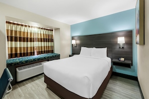 Una habitación de hotel con una cama grande, un área de descanso con un banco, un televisor montado en la pared y una mesita de noche con lámpara y teléfono.