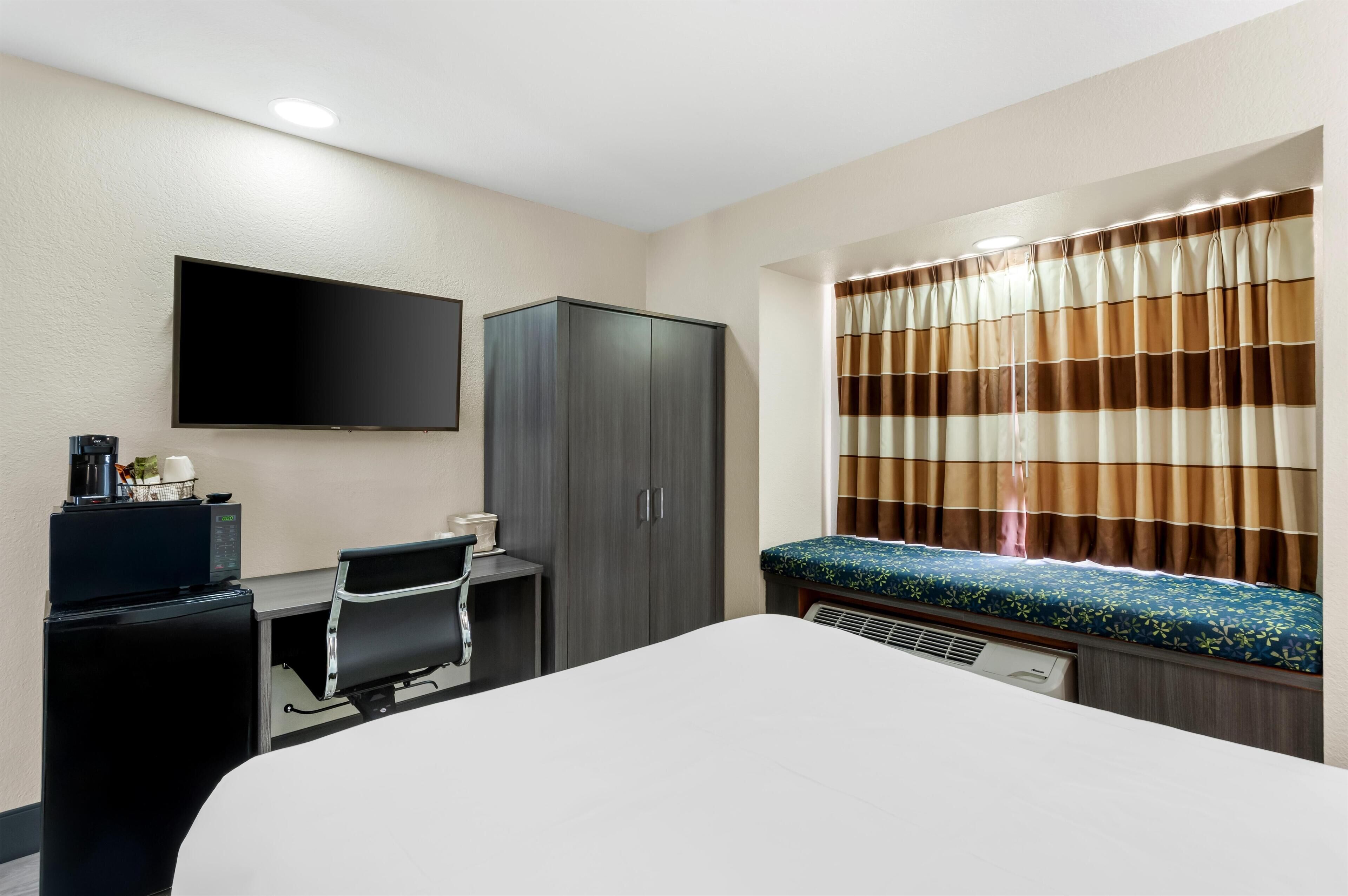 Habitación de hotel con cama, escritorio, silla, televisión, armario y ventana con cortinas.