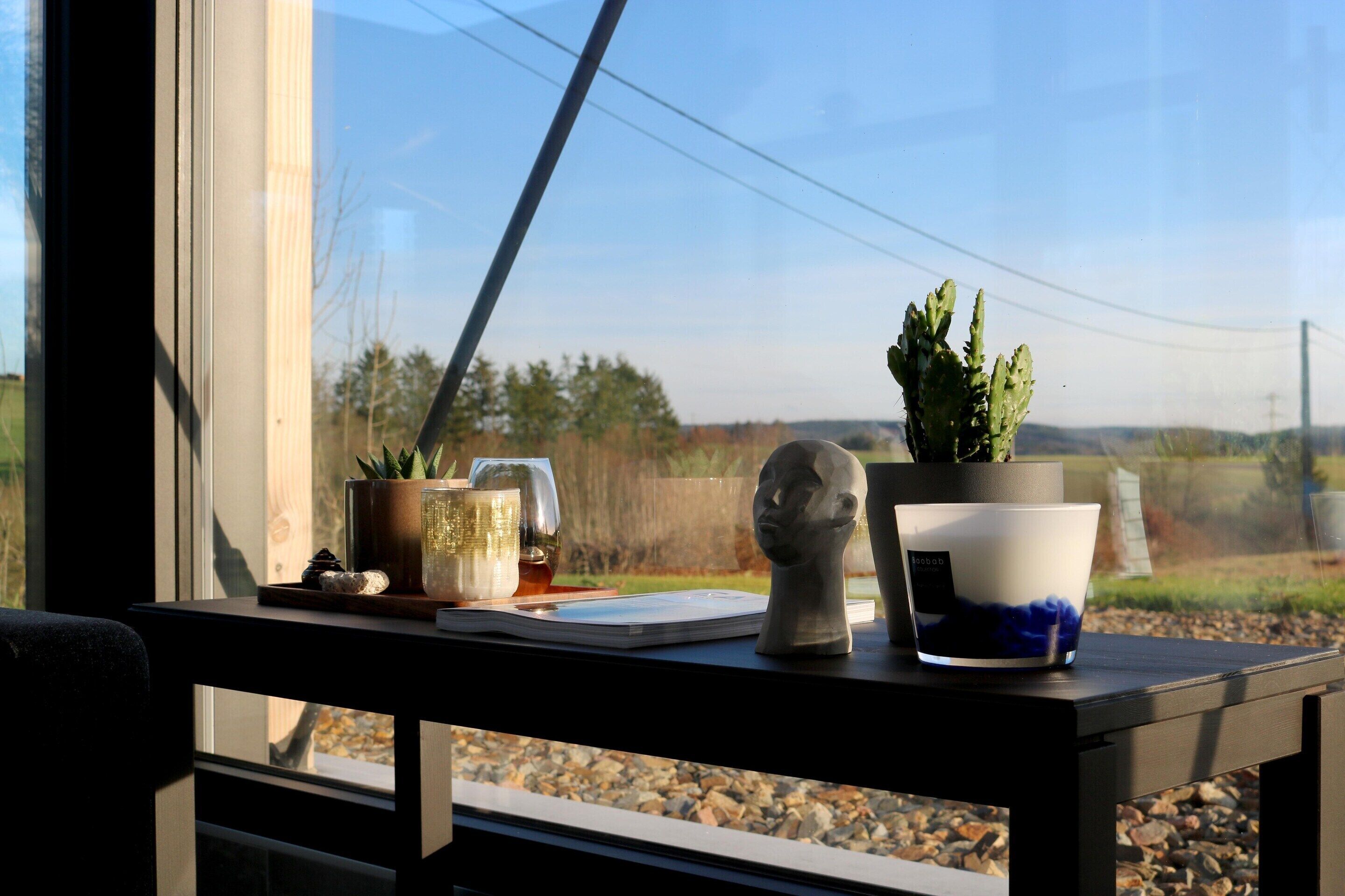 Une table avec un pot de fleurs, une tasse, un livre et un objet décoratif près d’une fenêtre.