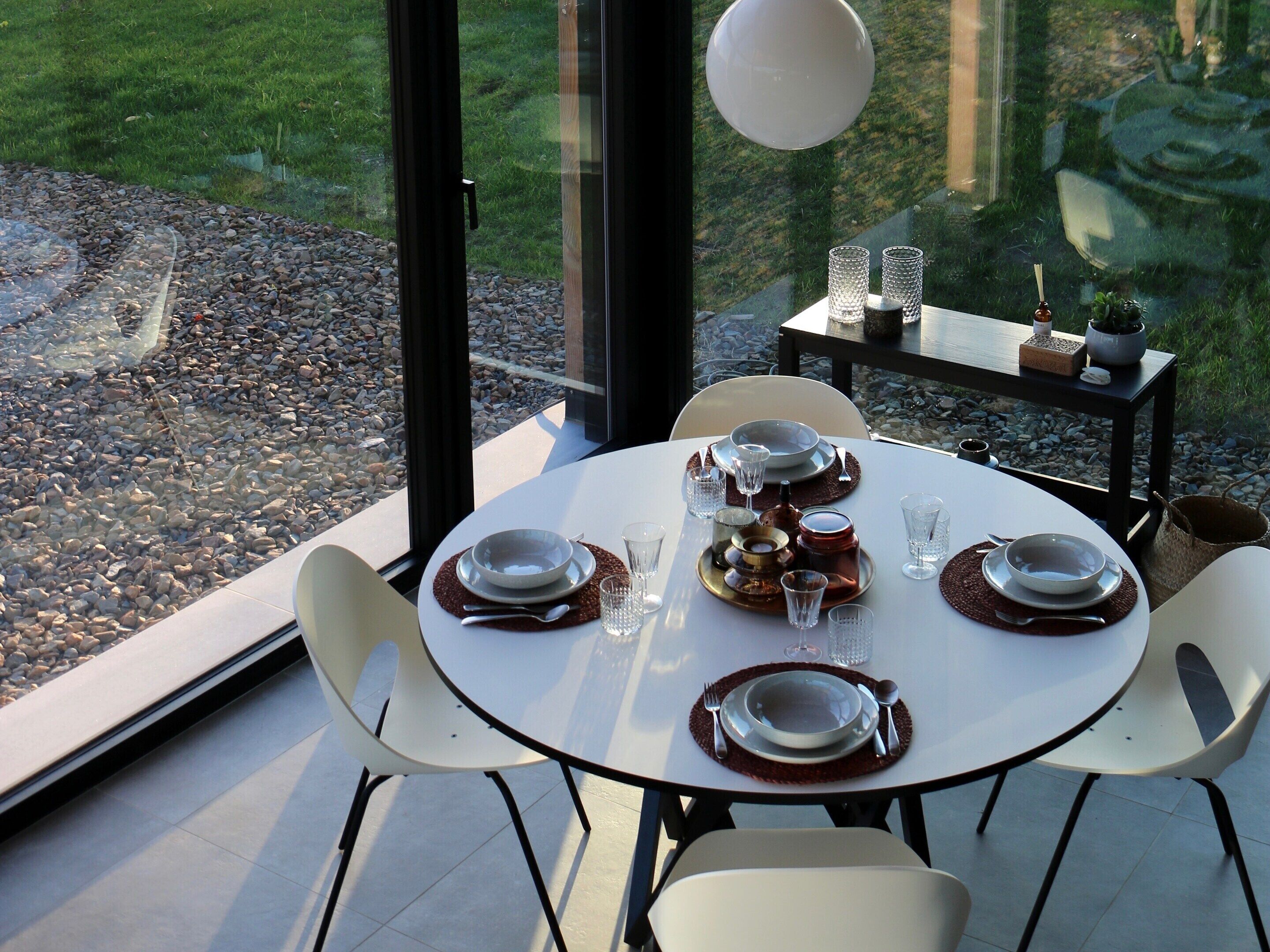 Ein Esstisch ist gedeckt mit Tellern, Gläsern und Besteck, umgeben von Stühlen, mit Blick auf einen Garten durch große Fenster.