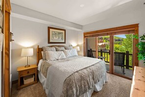 Ein Schlafzimmer mit einem großen Bett, einem Nachttisch, einer Lampe, einem gerahmten Bild und einer Glastür zum Balkon oder zur Terrasse, wo Tisch und Stühle stehen.