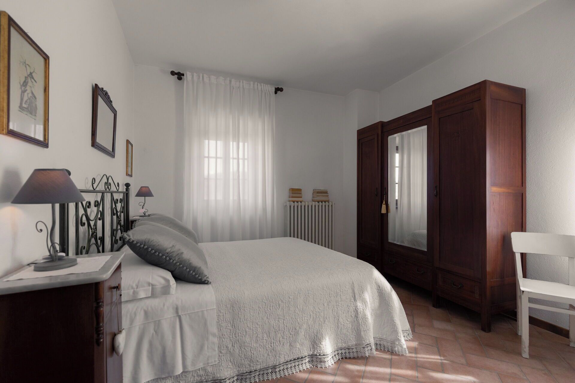 Une chambre à coucher comprenant un lit, une table de chevet, une lampe, une commode, une chaise et une fenêtre avec des rideaux.