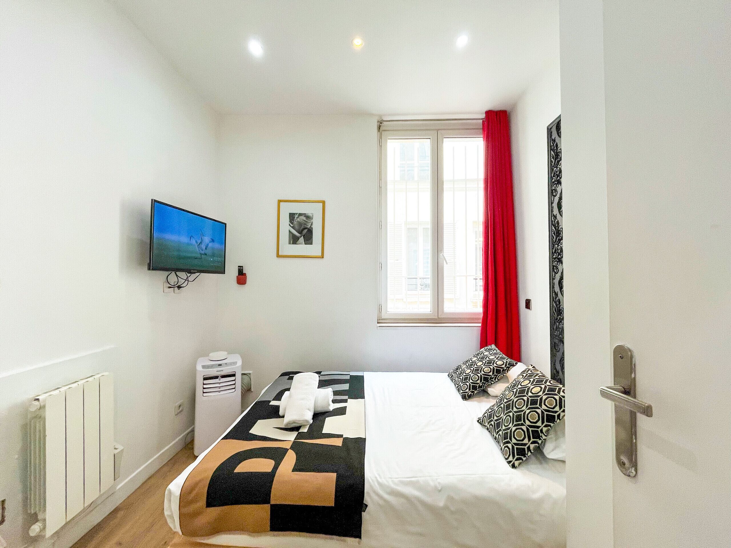 Une chambre d’hôtel avec un lit, une télévision, une fenêtre avec des rideaux rouges, un radiateur et un tableau accroché au mur.