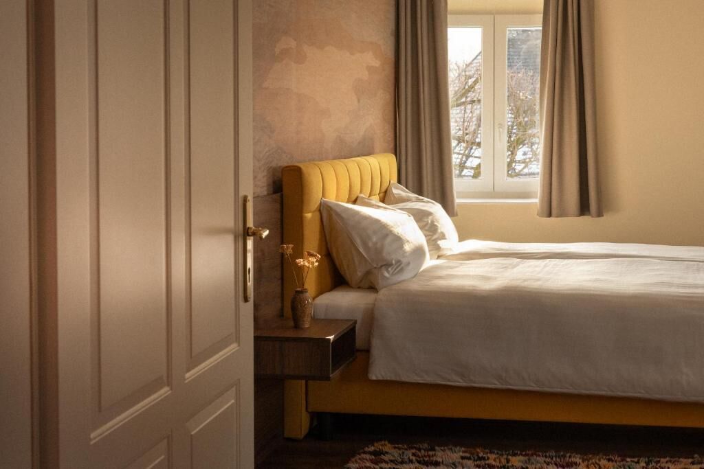 A bedroom with a bed, a nightstand, a vase with flowers, and a window with curtains.