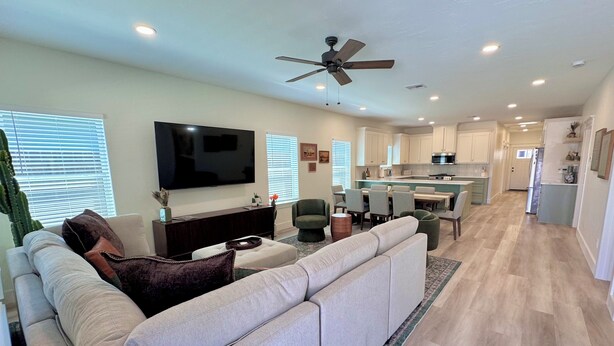 A modern living room with a large sectional sofa, a flat-screen TV, a ceiling fan, and a kitchen in the background. - ✪ Armadillo Rodeo ✪ 1 mile to Baylor, 1/2 mile to Magnolia Silos! (Waco)