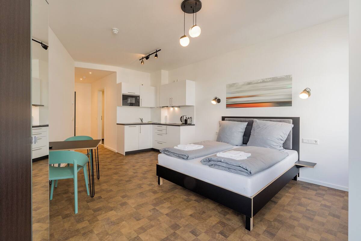 Un dormitorio moderno con cama, mesitas de noche, un escritorio y una silla. Cuenta con una kitchenette con armarios y un microondas.