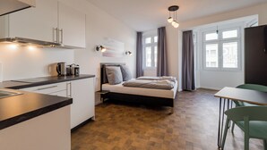 A compact hotel room with a kitchenette, a bed with two pillows, a bedside table, a window with curtains, and a green chair. - Nena Apartments Hermannplatz by LESA  (Berlin)