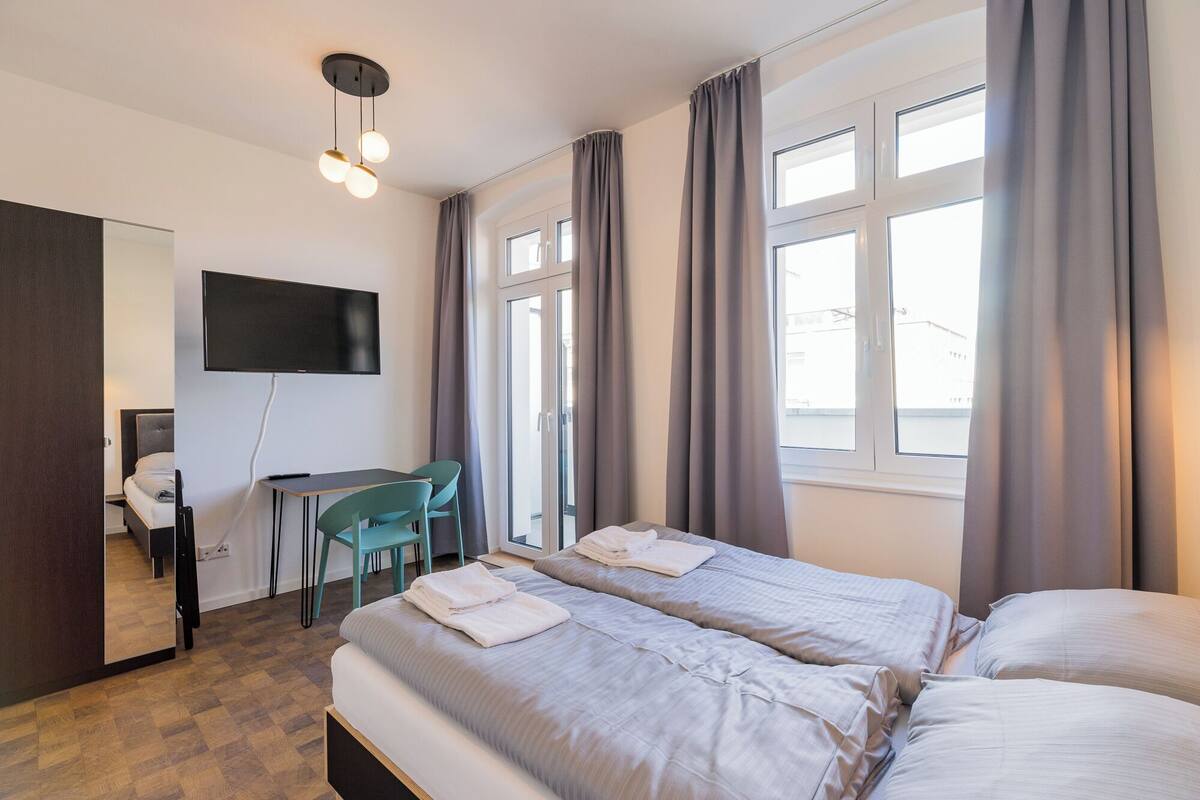 Una habitación de hotel con una cama, un escritorio, una silla, un televisor y ventanas amplias.