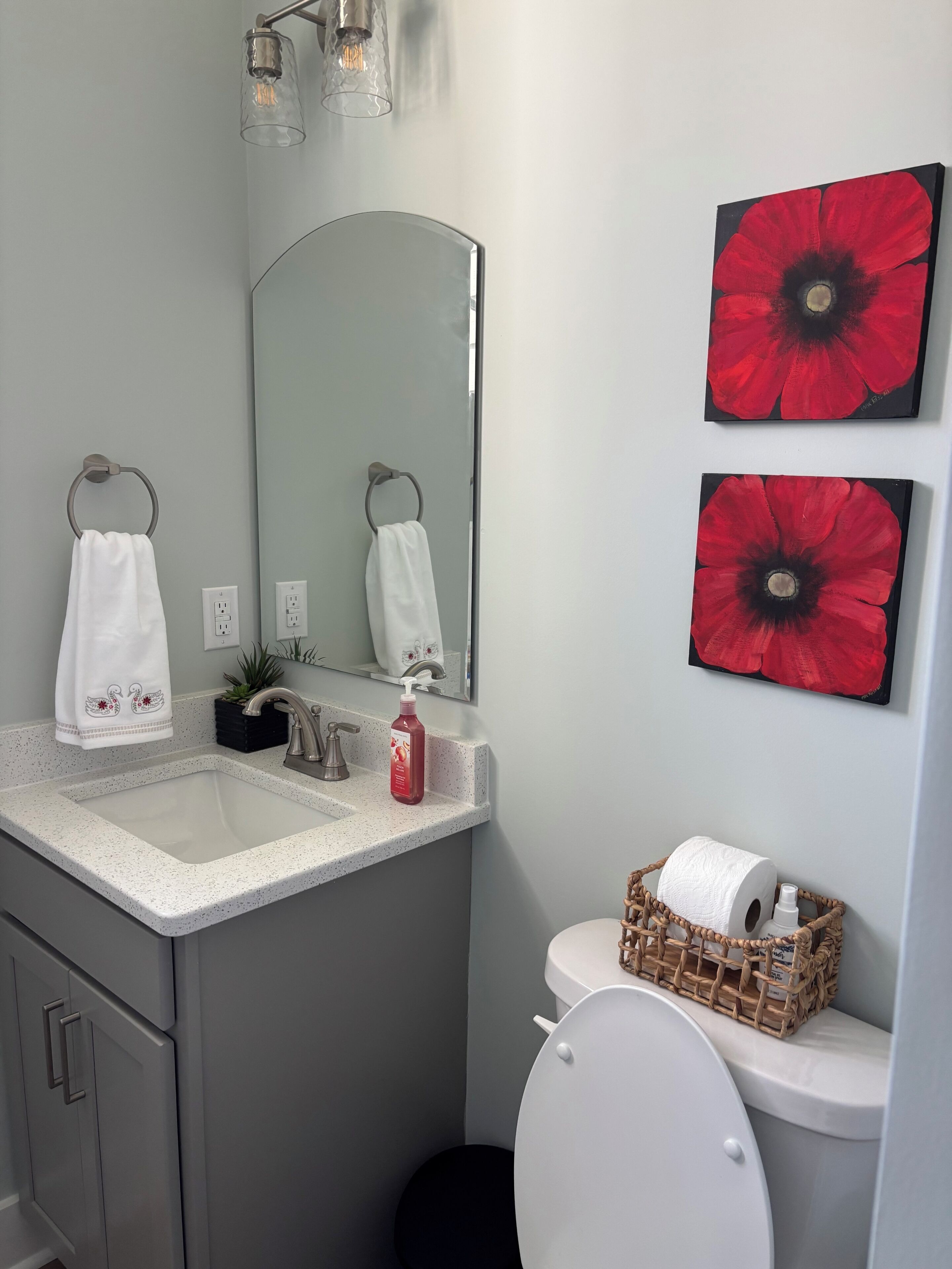 A bathroom with a white sink, two towels, a toilet, a mirror, and two red flower paintings.