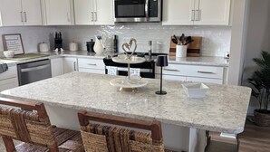 Une cuisine moderne avec des meubles blancs, un îlot central, deux chaises en osier et des luminaires suspendus.