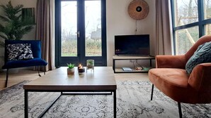 A living room with a sofa, a coffee table, a TV on a stand, a potted plant, and a large window. - Elevated Ewijk Cottage for 6 (Ewijk)