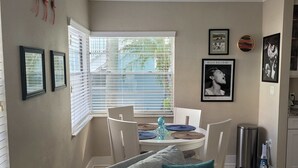 Un salon avec un canapé gris, des coussins turquoise, un coin repas avec une table et des chaises, et un coin cuisine avec une poubelle et une bouilloire.