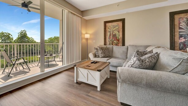 A spacious living room with a balcony, a ceiling fan, a sofa, a coffee table, and a painting on the wall. - Mangrove View 2BR - Pool Hot Tub Gym Access (Ruskin)
