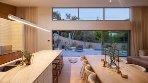A modern kitchen with a large island, a dining area with a wooden table and chairs, and a view of a backyard with a wooden deck and outdoor furniture. - Bridgewater House Gas Heated Pool (Melbourne)