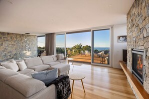 A modern living room with a stone wall, a large sofa, a fireplace, and a view of the outdoors. - Airosa Blanca Pool Spa Sea Views (Melbourne)