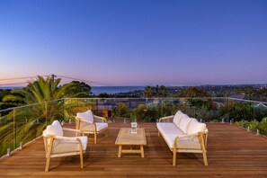 Um deck de madeira com móveis de vime e uma mesa de vidro, com vista para um horizonte urbano ao entardecer.