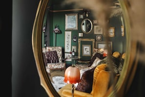 Quarto com paredes verdes, um espelho redondo, quadros emoldurados, uma mesa com uma garrafa e duas cadeiras estampadas.