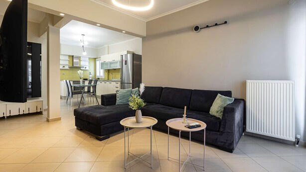 A modern living room with a black sofa, two small round tables, a TV, and a kitchen in the background. - Faliro Escape - Spacious Stay in Thessaloniki (Thessaloniki)
